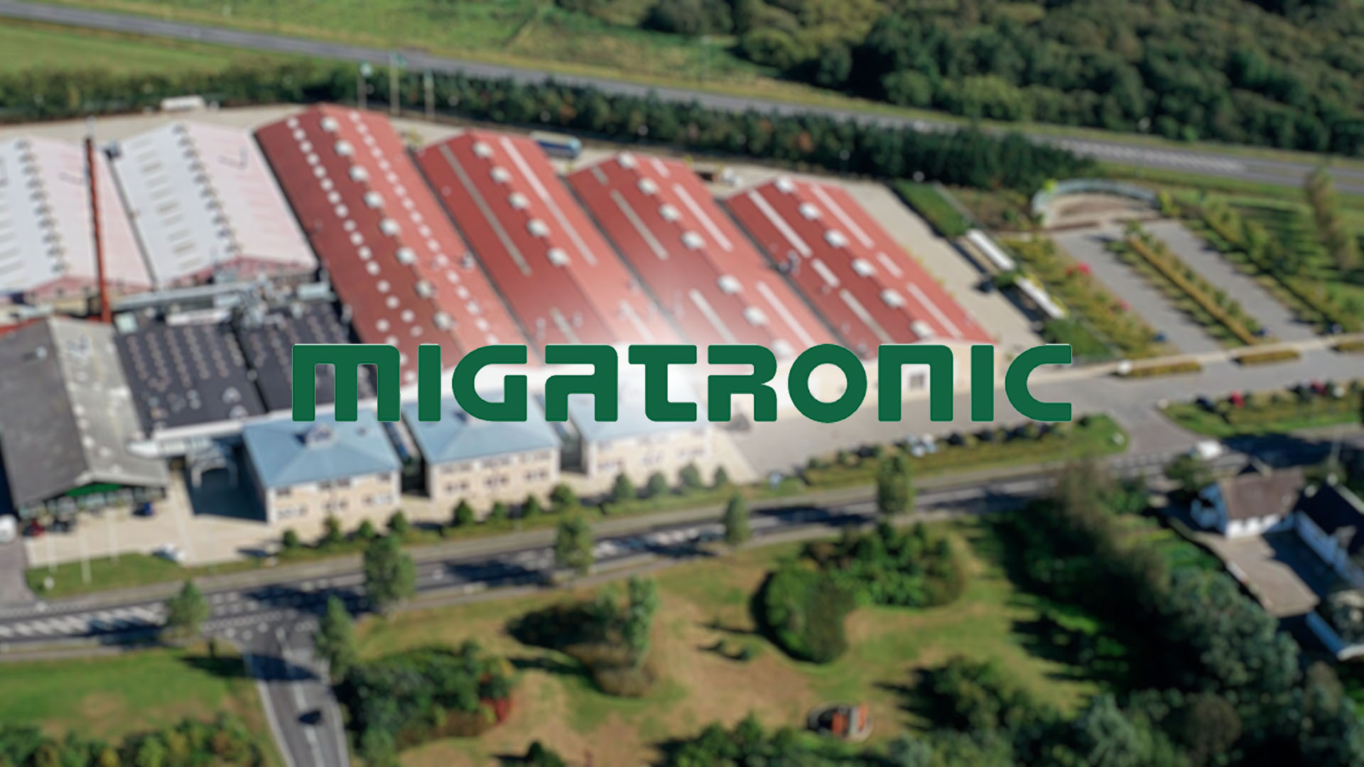 Aerial view of a large industrial complex with multiple red-roofed buildings surrounded by greenery. The logo "MIGATRONIC" is prominently displayed.