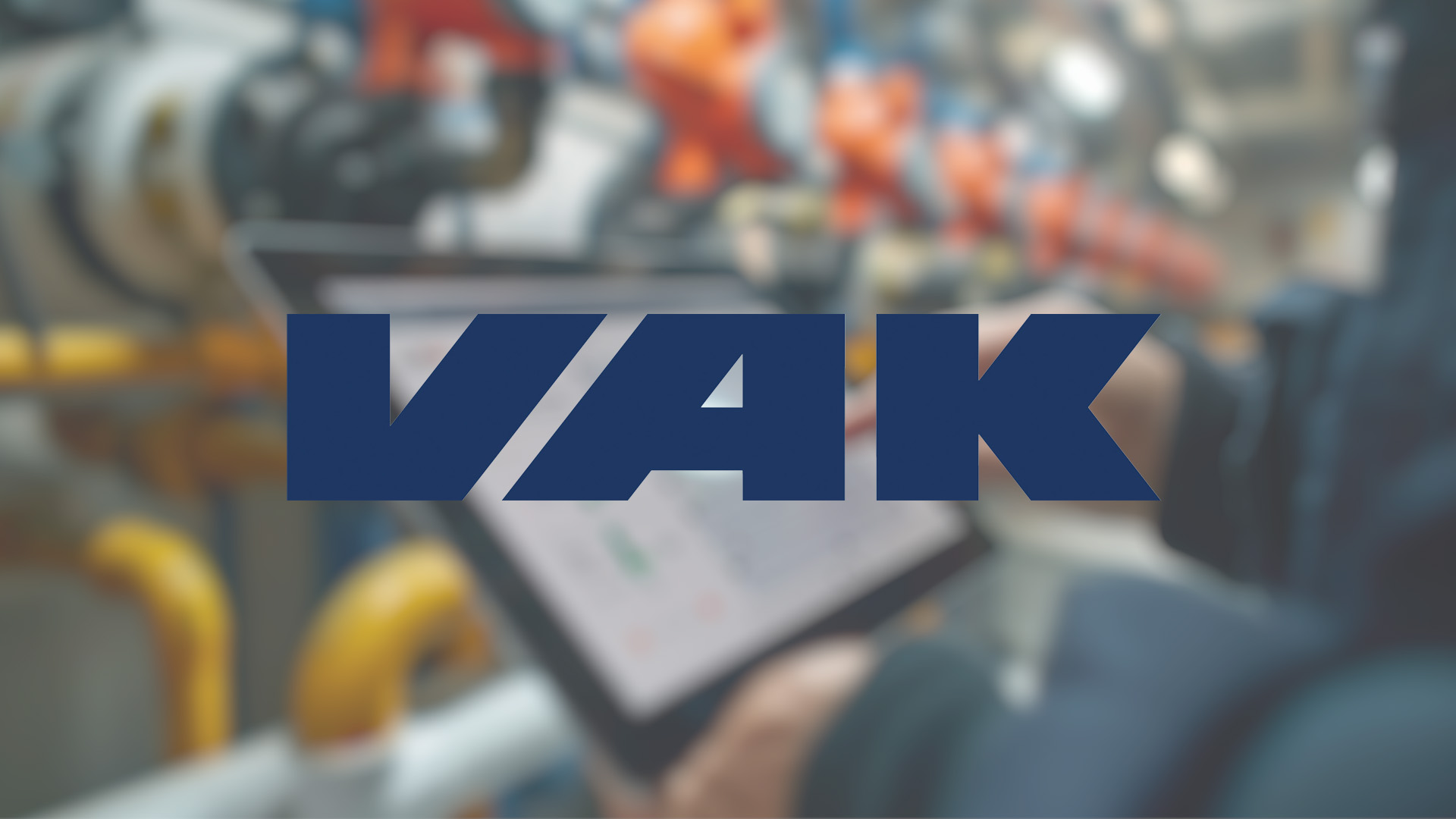 Blurred background of industrial pipes with a person using a tablet. Large bold “VAK” logo is overlaid in the center.