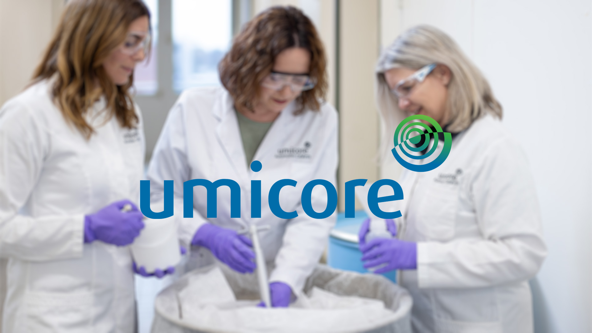 Three women in lab coats and safety goggles work together in a lab, wearing purple gloves. The blurred Umicore logo overlays the image.